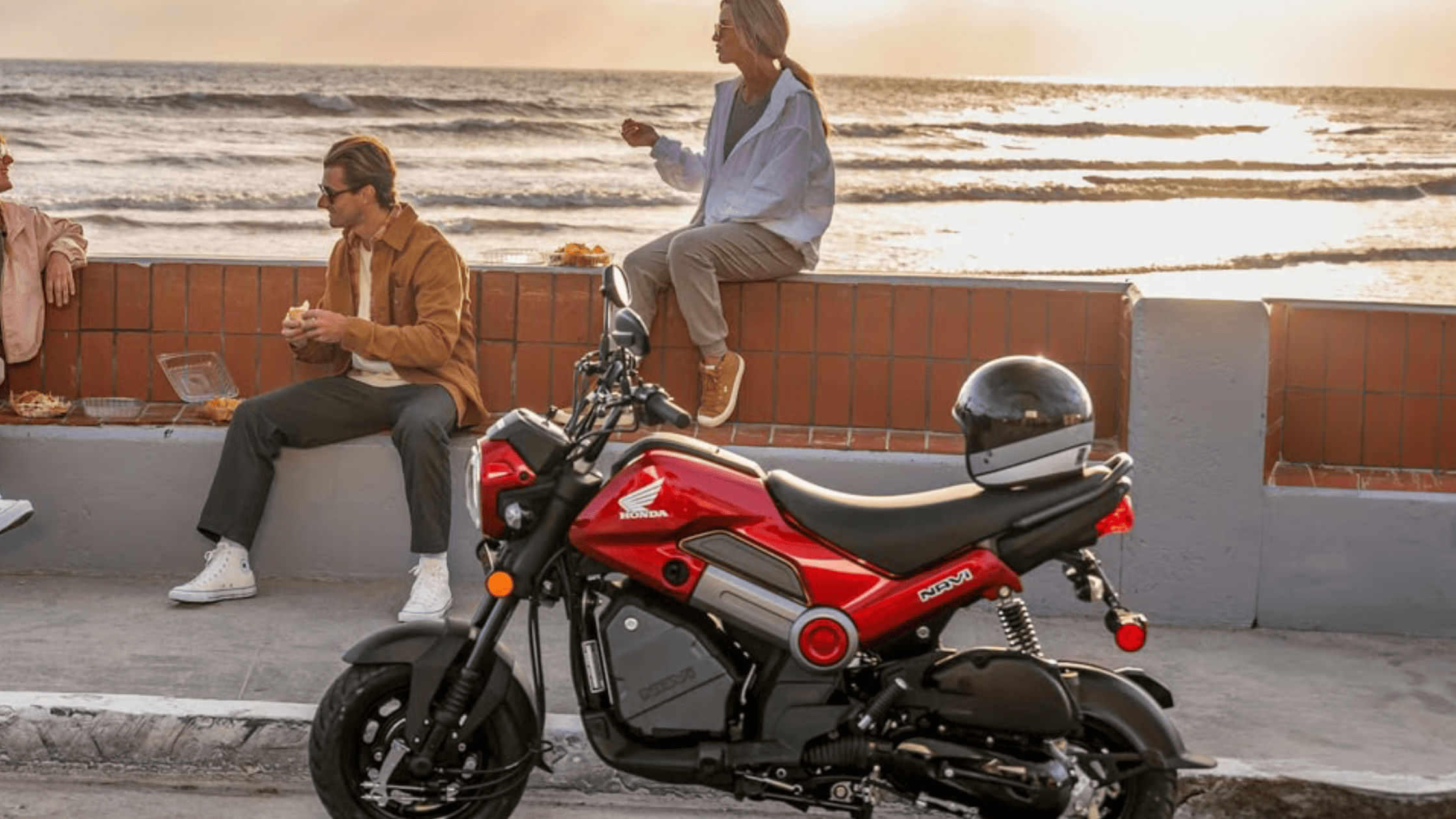 Red 2025 Honda Navi parked near ocean boardwalk while friends enjoy sunset by the water.