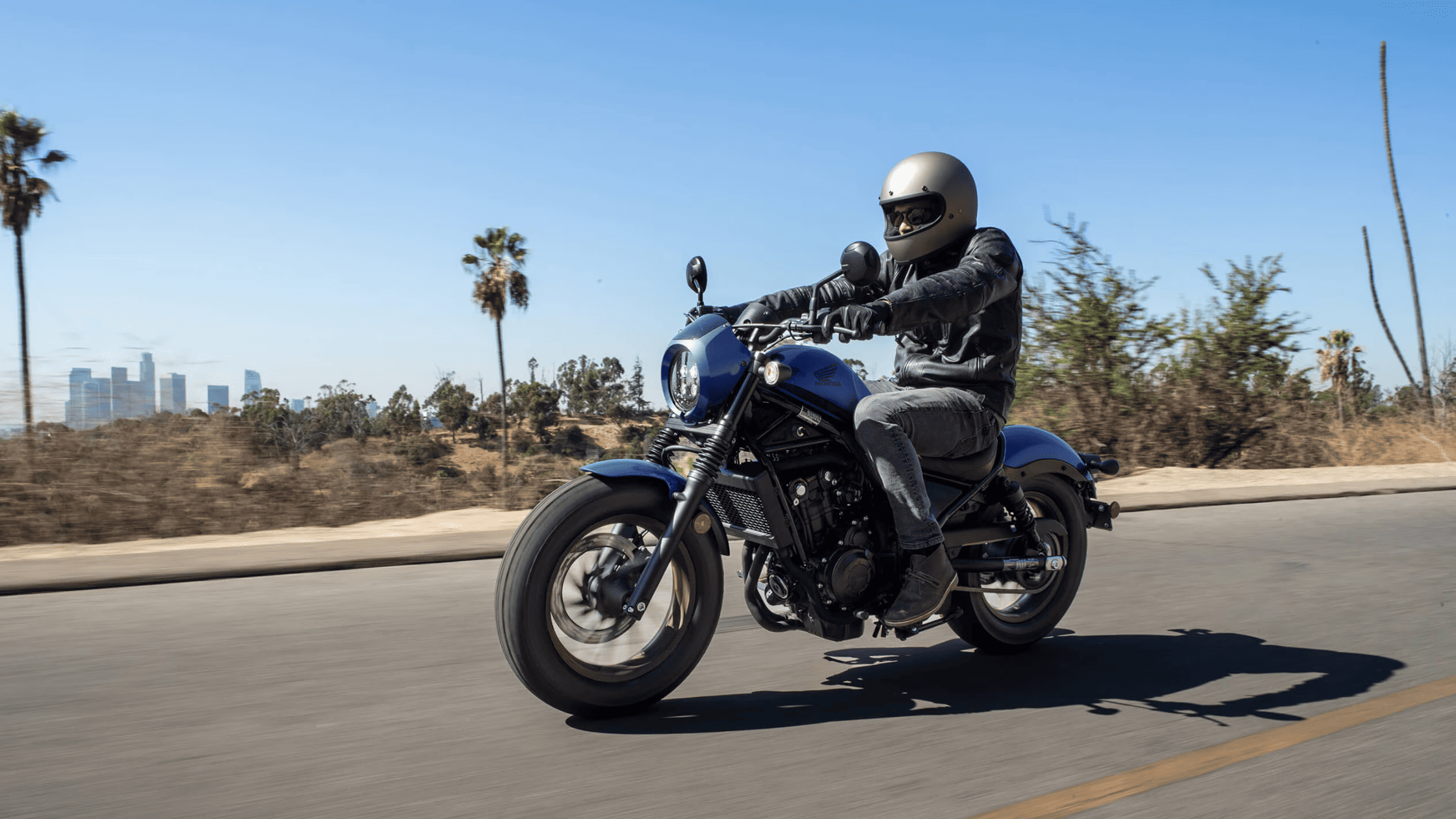 Woman on matte black 2025 Honda Rebel 1100 DCT SE motorcycle cruising through urban setting
