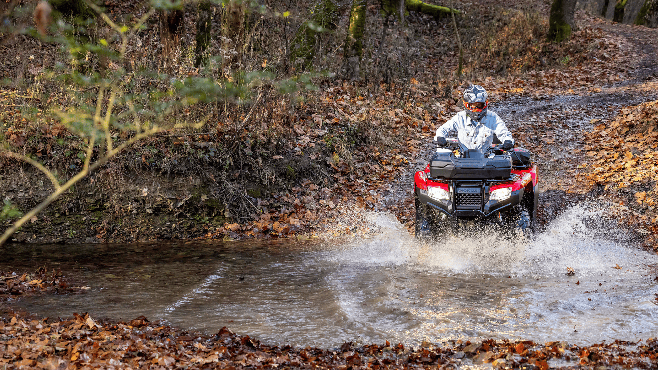 Red Honda FourTrax Rancher 4x4 ATV crossing water, rider in helmet, forest trail
